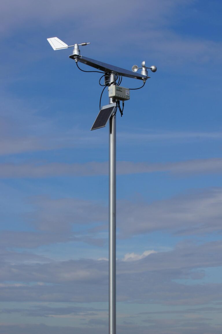 Anemometer and weather vane atop a tall pole under a clear blue sky.